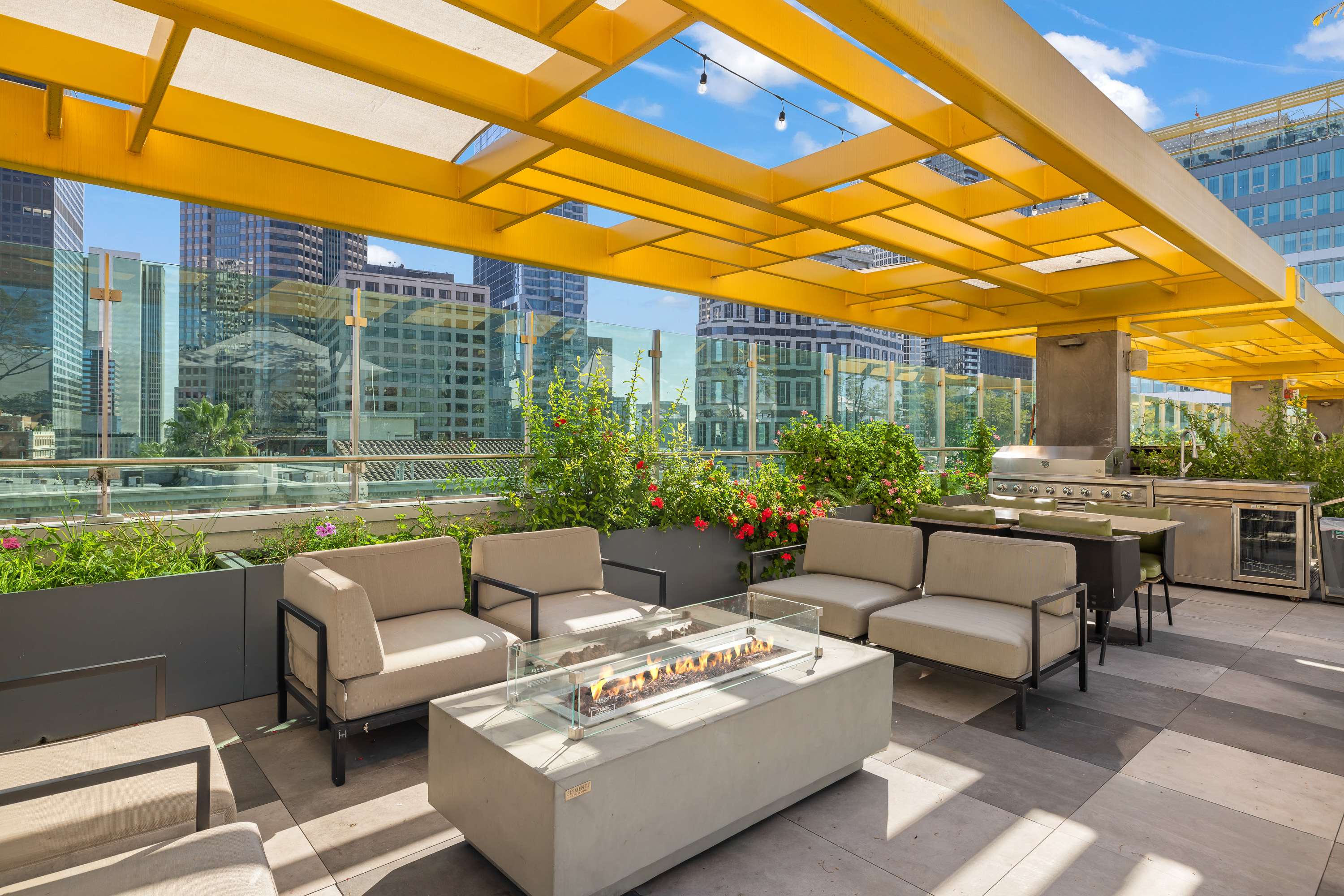 Rooftop lounge at Skylux on Wilshire with cushioned seating, a fire table, barbecue grills, landscaped planters, and Downtown Los Angeles views.